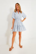 A woman smiles while wearing a light blue Hyacinth House Ditsy Loren cotton floral mini dress with tan sandals, holding a white bottle.