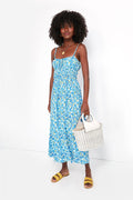 A woman in a FAITHFULL THE BRAND blue floral summer midi dress, yellow sandals, and holding a white woven handbag stands confidently.
