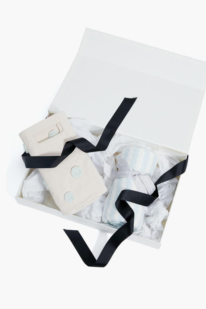 The Tuckernuck Tots white gift box with black ribbon holds a folded cream cloth and a rolled blue-and-white striped towel on white tissue, making it an adorable option for baby gifts or a thoughtful nursery bundle.