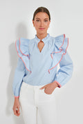 A woman wears the Hyacinth House Holly Blouse, a blue and white striped, feminine blouse with ruffle detail and red trim, styled with white pants.