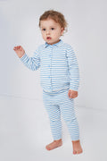 A toddler stands barefoot in a bright room, wearing a Rachel Riley British children’s knitwear two-piece cotton set: a blue and white striped knit cardigan and matching pants, gazing slightly to the side.