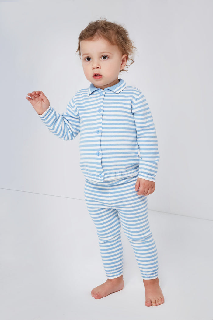 A toddler stands barefoot in a bright room, wearing a Rachel Riley British children’s knitwear two-piece cotton set: a blue and white striped knit cardigan and matching pants, gazing slightly to the side.