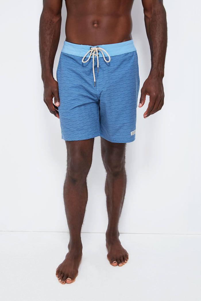 A man stands barefoot in Fair Harbor blue boardshorts, featuring quick-drying fabric, a light blue waistband, and a drawstring tie.
