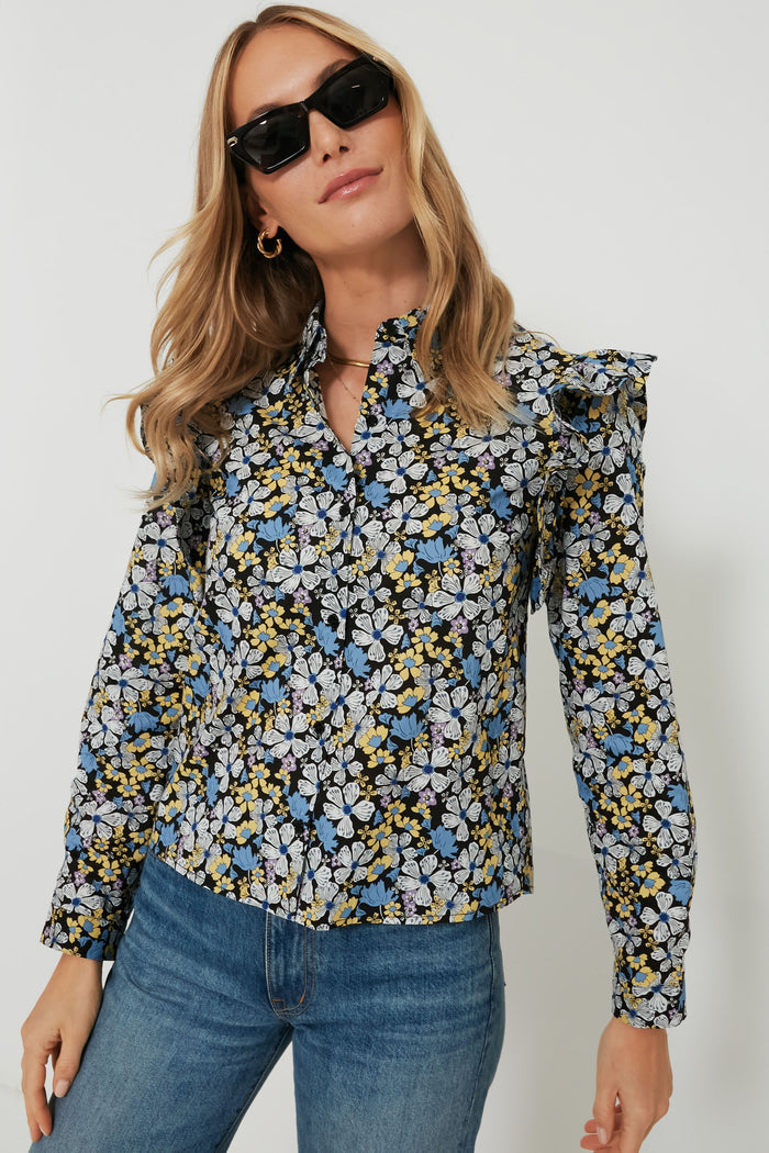 A woman stands confidently, wearing Hyacinth House sunglasses and a long-sleeve floral blouse with ruffle details, paired with blue jeans.
