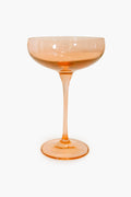 A single empty peach-colored coupe glass from Estelle Colored Glass, featuring hand-blown colored glass with a wide bowl and slender stem, exemplifies luxury stemware.