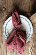 A Madame de la Maison white plate with a blue patterned rim displays a folded maroon linen napkin tied with twine on a rustic wooden table—an elegant, sustainable tableware choice.