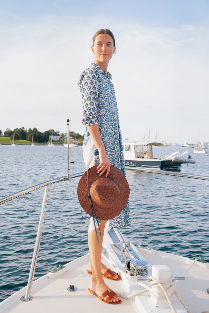 A woman in a blue patterned dress stands on a boat deck, holding an oversized fedora by Lola Hats, with water and boats in the background.