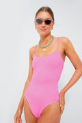 A woman models the Hunza G pink crinkle-stretch one-piece swimsuit with a classic scoop neckline, sunglasses, and layered necklaces, highlighting its high-cut style.