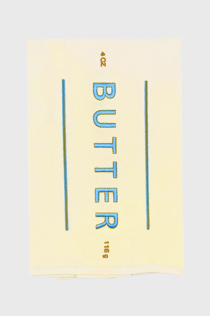 A Lynen Butter Me Up Tea Towel displays a stick of butter labeled "BUTTER" in blue, with 4 OZ and 113g in brown—an embroidered cotton kitchen towel that's perfect for your kitchen.