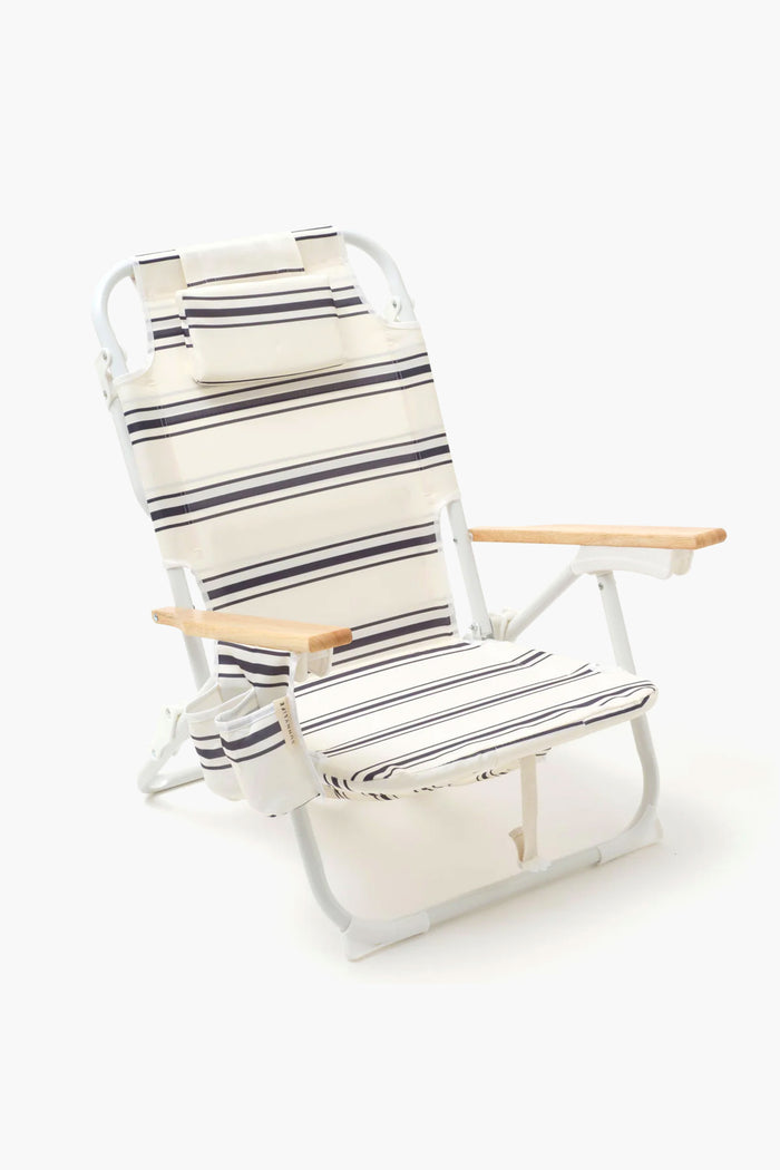 The Sunnylife foldable beach chair features white and navy stripes, wooden armrests, a headrest, and a side pocket—combining comfort and style for relaxing by the sea.
