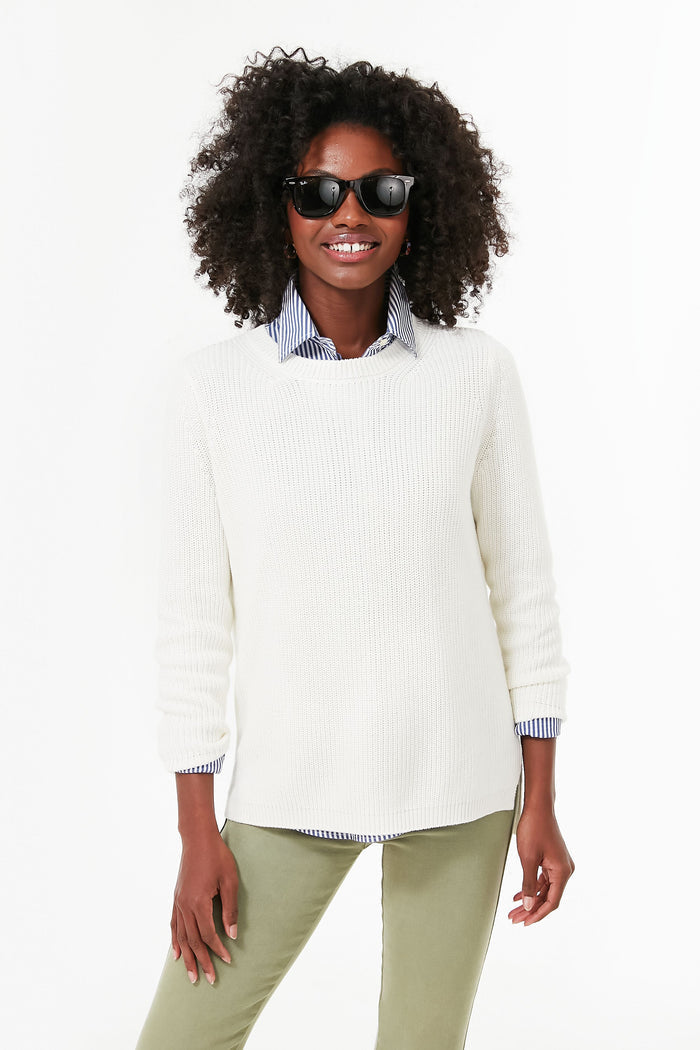 A smiling woman with curly hair wears 525 America sunglasses, a cotton crewneck pullover over a striped shirt, and light green pants.