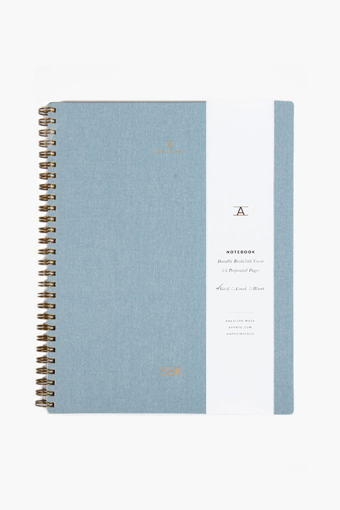 The APPOINTED Chambray Blue Lined Notebook features a spiral binding, white vertical label with gold text and accents, a durable book-cloth cover, and recycled paper.