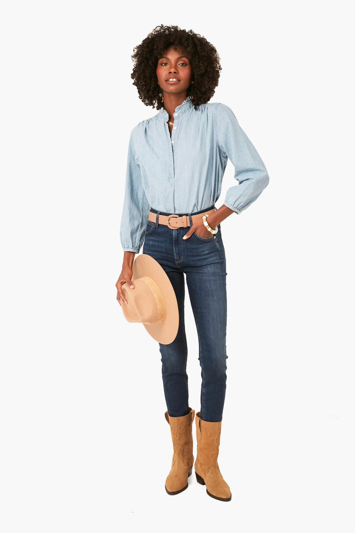 A woman wearing a TROVATA Birds of Paradis Chambray Henley with a ruffle collar, dark blue jeans, tan belt and boots, holds a tan hat at her side.