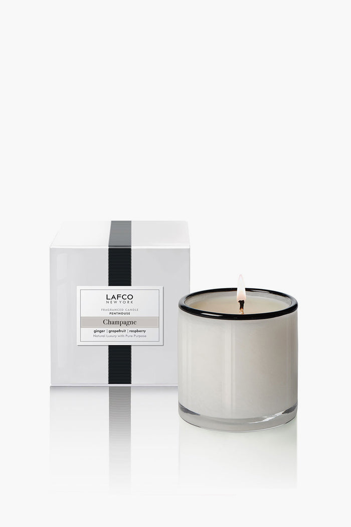 A white candle in a clear glass jar is lit beside its LAFCO New York Champagne Signature Candle box—a luxurious home fragrance and perfect housewarming gift.