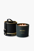 A black Sicily Hill Champagne Candle with three flames sits next to its matching black and gold leather box, both exuding elegance.