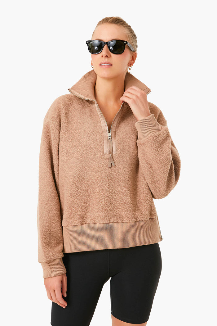 A woman wears black sunglasses, a tan Varley half zip fleece pullover made from recycled borg fleece, and black shorts, creating a stylish layered look.
