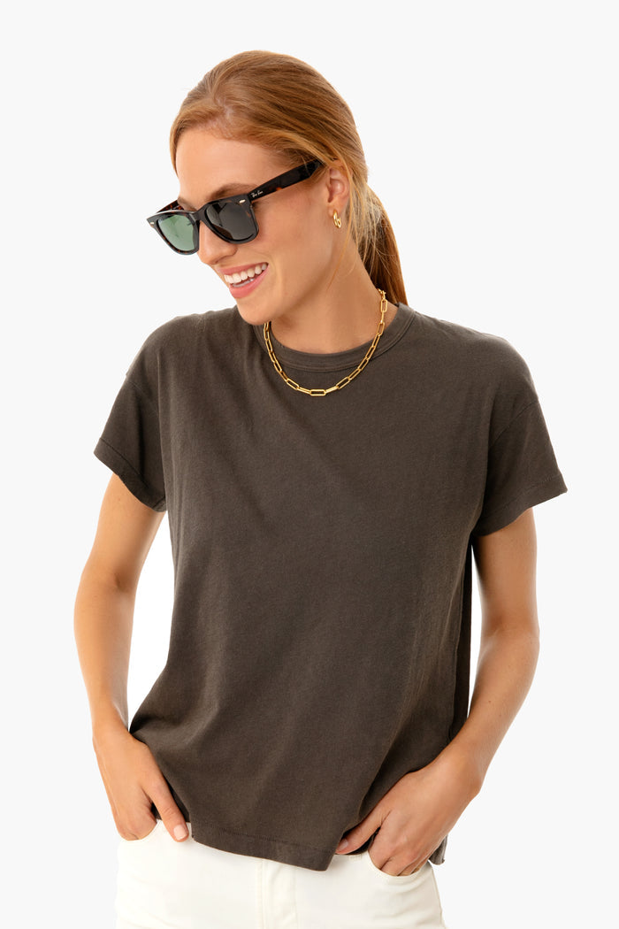 A woman stands confidently wearing black sunglasses, a gold chain necklace, white pants, and THE GREAT. dark grey 100% Cotton Boxy Crew Tee.
