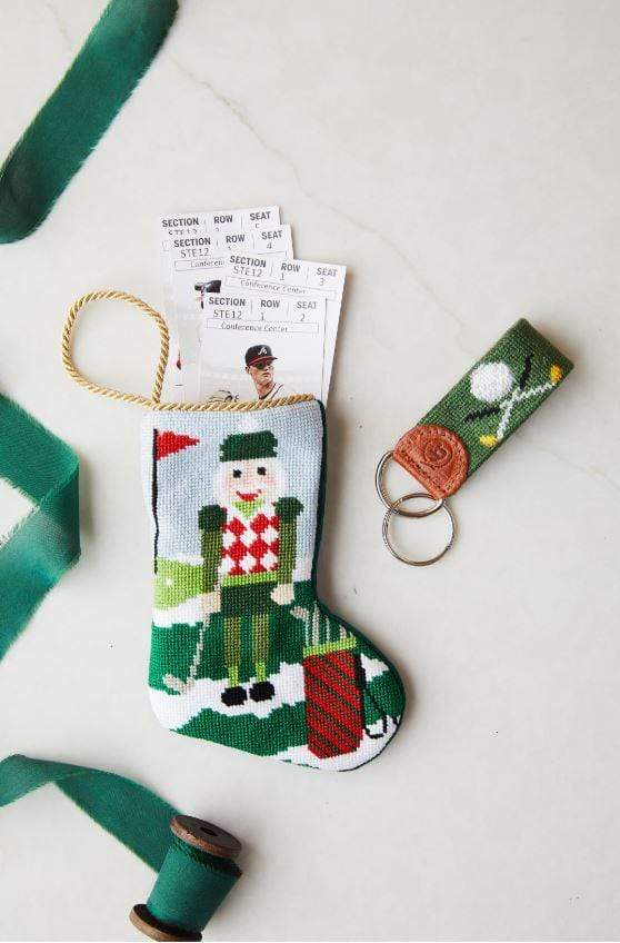 A small, hand-stitched Bauble Stockings Christmas stocking with a golfer design holds event tickets. Next to it are a green and brown golf-themed keychain and a spool of green ribbon from Bauble Stockings.
