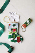 A small, hand-stitched Bauble Stockings Christmas stocking with a golfer design holds event tickets. Next to it are a green and brown golf-themed keychain and a spool of green ribbon from Bauble Stockings.