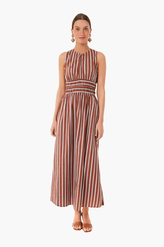 A woman wears a Faithfull sleeveless, ankle-length brown and white vertically striped handmade cotton dress with a gathered waist—perfect for vacation.