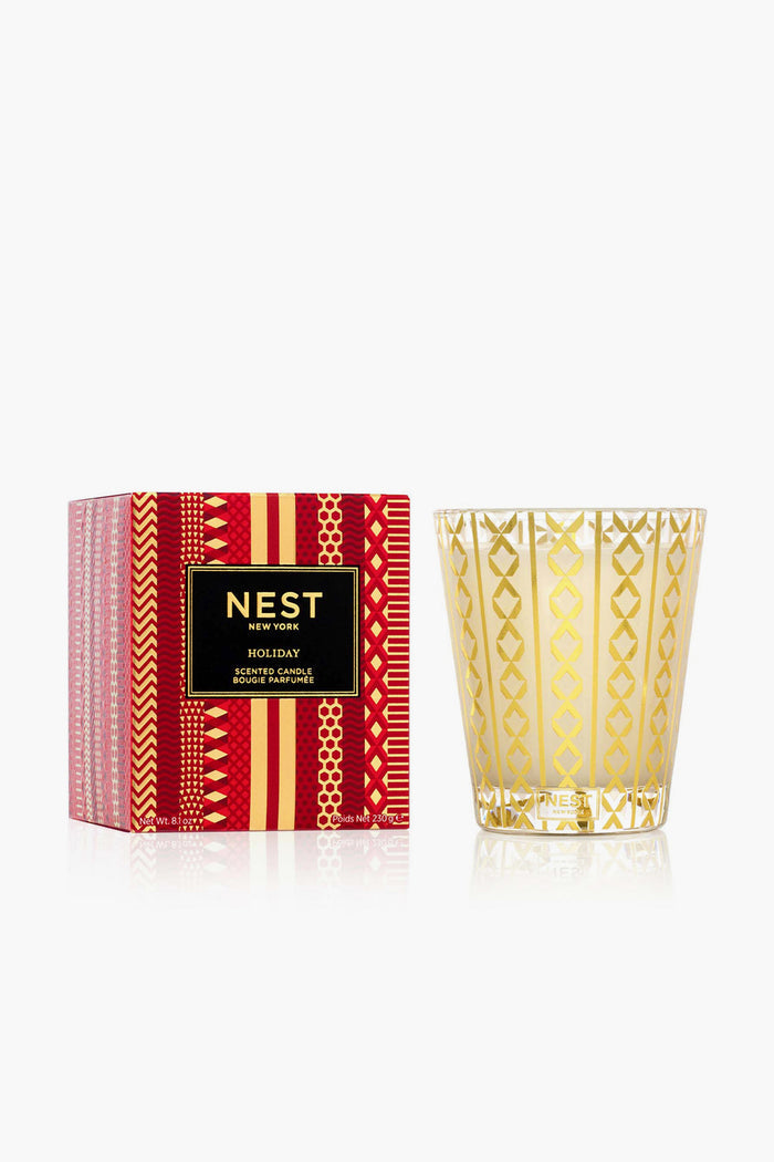 A NEST New York holiday soy wax candle in a decorative gold-patterned glass is displayed beside its festive red and gold box.