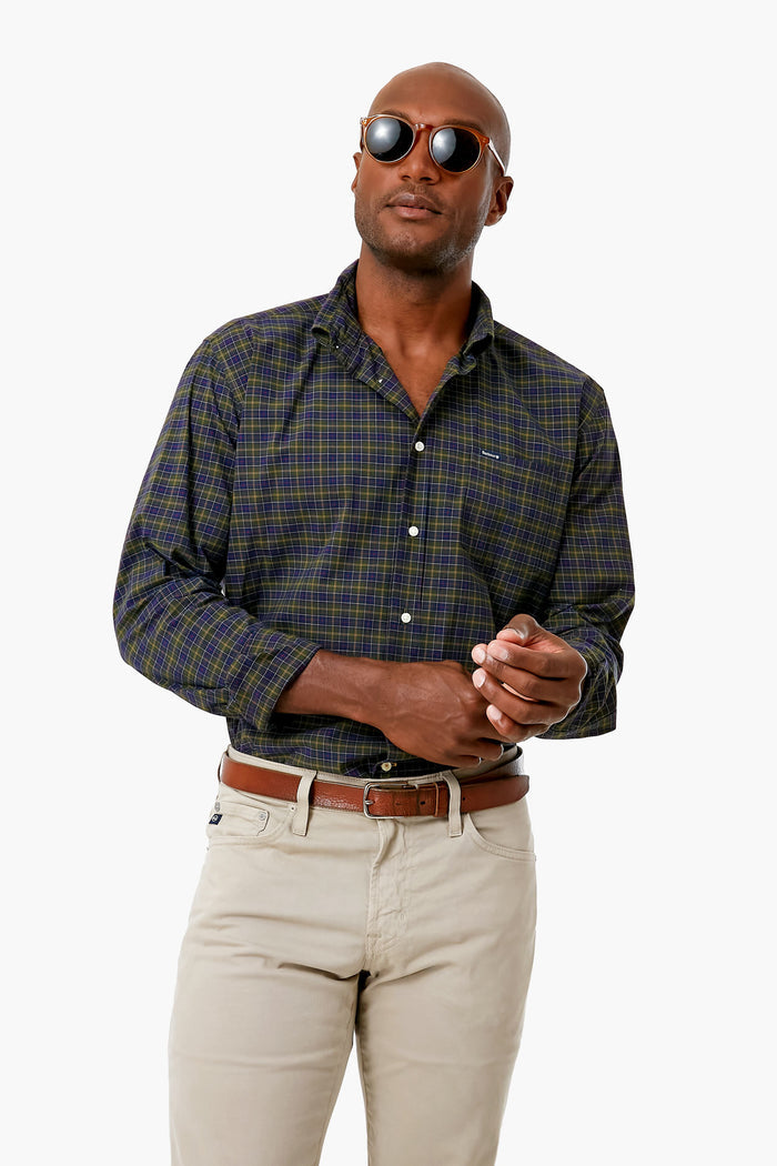 A man wearing sunglasses, a dark Barbour tartan button-up shirt, and beige pants stands confidently.