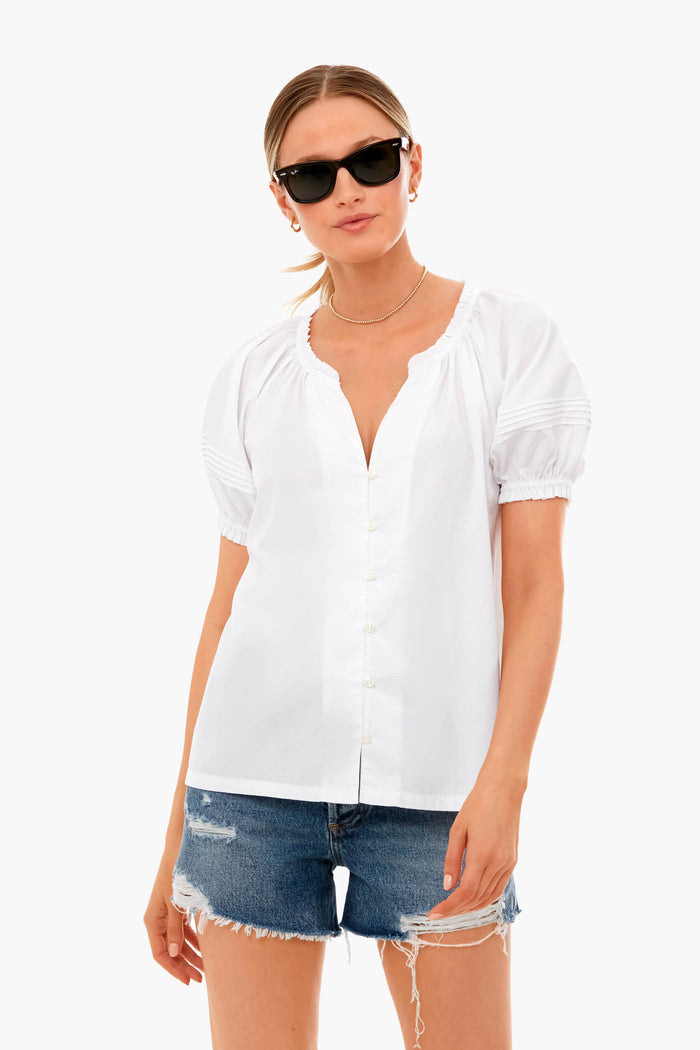 A woman wears a TROVATA Birds of Paradis Classic White June Blouse with a ruffle collar, denim shorts, and sunglasses, highlighting this lightweight cotton summer blouse.