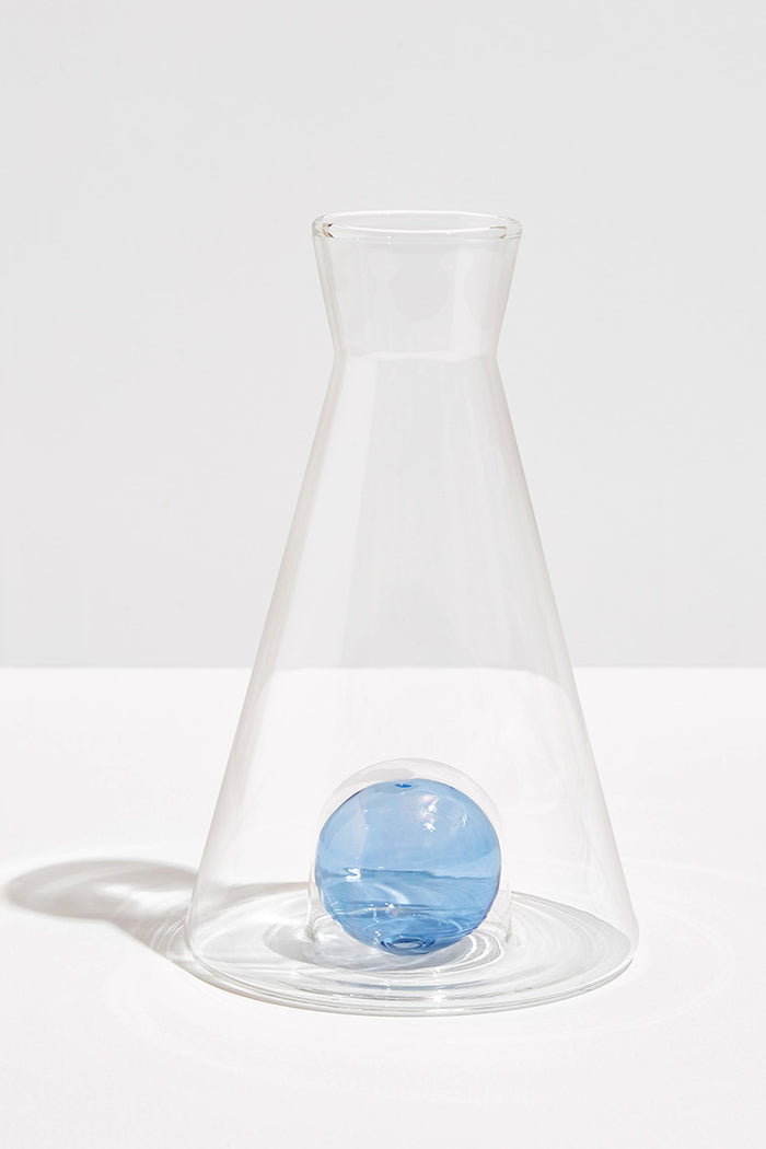 The FAZEEK clear borosilicate glass flask features a wide base, narrow neck, and a single blue glass sphere at the bottom for an elegant sculptural homeware accent.