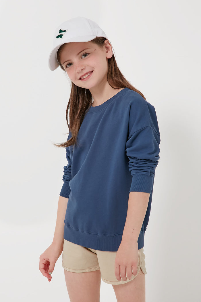 A girl in Les Gamins' organic cotton sweatshirt, beige shorts, and white cap smiles at the camera—stylish and sustainably made kids' clothing that’s comfortable too.