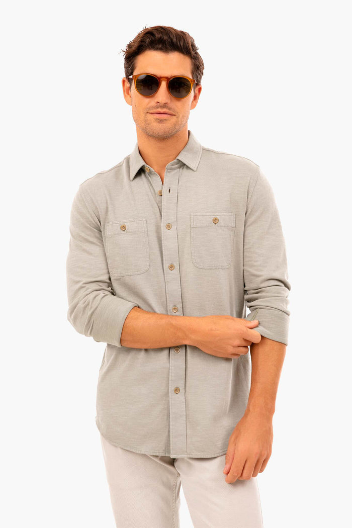 A man wearing sunglasses, light pants, and a Faherty light gray knit seasons shirt with two chest pockets made from organic cotton stands confidently.