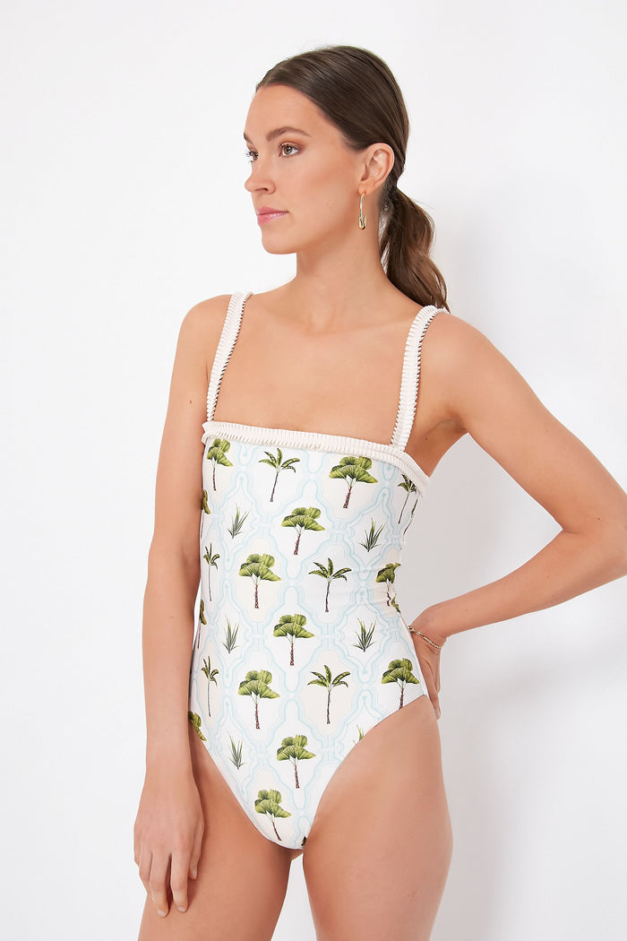 A woman stands confidently with her hand on her hip, wearing an AGUA BY AGUA BENDITA one-piece swimsuit featuring tropical palm tree and plant prints.