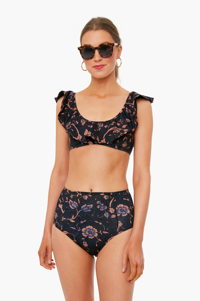 A woman in sunglasses and hoop earrings models a Ulla Johnson black floral bikini featuring a ruffled neckline and high-waisted bottoms.