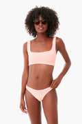 A woman stands confidently in the light, wearing a Hunza G light pink crinkle-stretch bikini with a scoop neckline and round sunglasses.