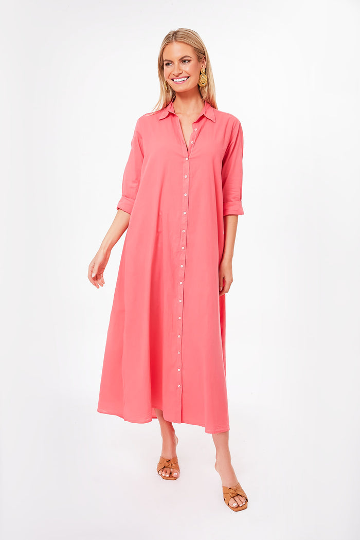 A woman stands wearing a coral pink cotton poplin dress with rolled-up sleeves from XiRENA, tan sandals, and gold earrings.
