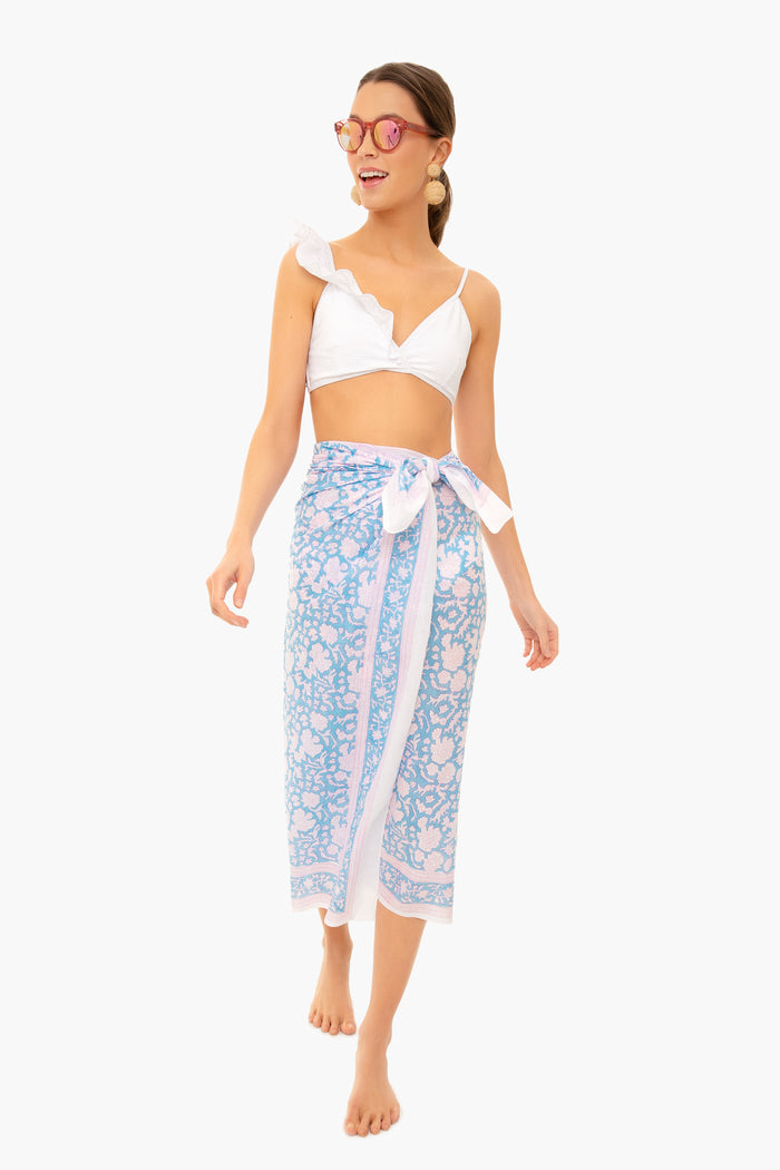 A woman in a white bikini top, pink sunglasses, and an SZ Blockprints light blue and pink hand block printed cotton Indian sarong as a beach coverup stands barefoot.