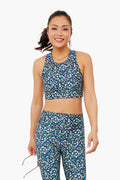 A woman wearing a blue floral Addison Bay sweat-wicking activewear set with a long line sports bra holds glasses in one hand and smiles.