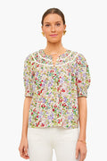 A woman faces forward, wearing THE GREAT. Storyteller Top—a floral blouse with lace trim—and white pants. Her hair is tied back, and she accessorizes with gold earrings, a necklace, and a bracelet.