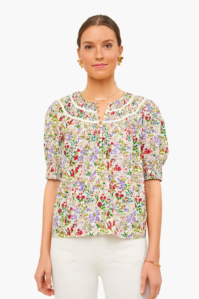 A woman faces forward, wearing THE GREAT. Storyteller Top—a floral blouse with lace trim—and white pants. Her hair is tied back, and she accessorizes with gold earrings, a necklace, and a bracelet.