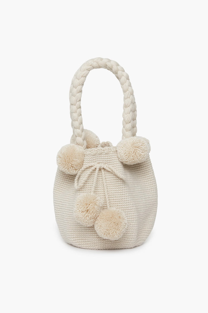 A cream-colored handwoven bag by Marina Orth Foundation from San Jacinto, Colombia, featuring braided handles and decorative pom-poms.