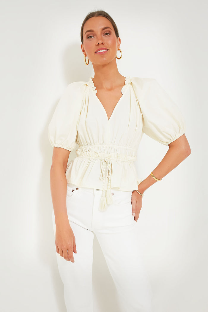 A woman wears a Moon River cream peplum top with puff sleeves and a drawstring waist, paired with white pants. She accessorizes with hoop earrings and gold jewelry.