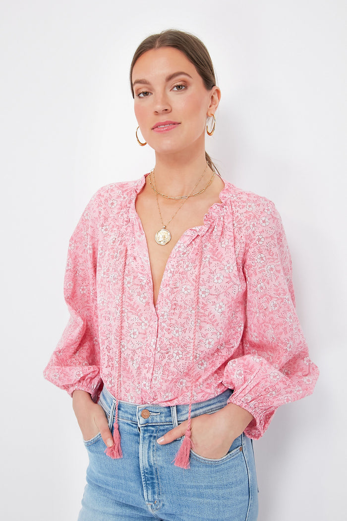 A woman wears a pink floral organic cotton blouse with oversized blouson sleeves from Love Tomas by Emerson Fry, paired with light blue jeans, gold hoop earrings, and layered necklaces for a comfortable and stylish look.