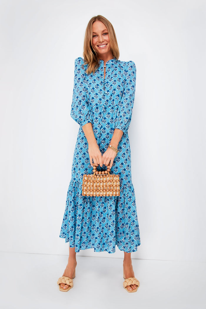 Woman in a blue floral boho dress from Love Tomas by Emerson Fry, carrying a small woven handbag and wearing beige sandals.