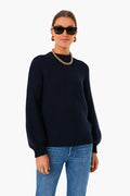A woman stands confidently in a Dark Navy Button Back Crewneck Sweater by Alex Mill, blue jeans, a gold chain necklace, hoop earrings, and black sunglasses.