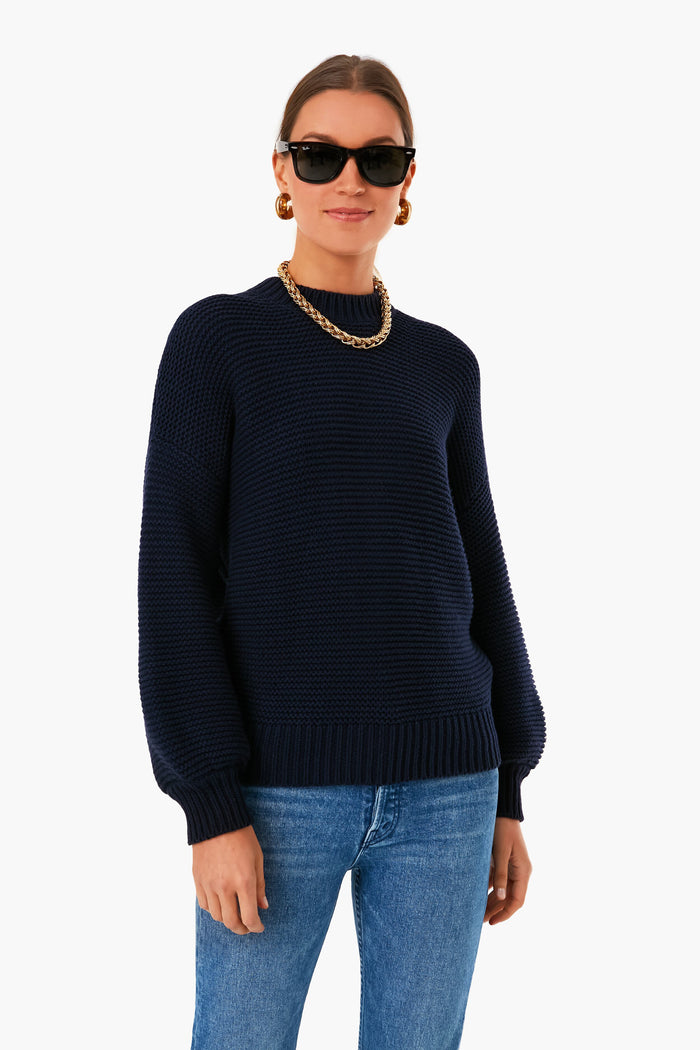 A woman stands confidently in a Dark Navy Button Back Crewneck Sweater by Alex Mill, blue jeans, a gold chain necklace, hoop earrings, and black sunglasses.