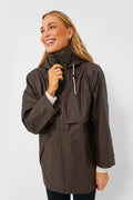 A woman wearing a Varley Alyssa Rain Jacket, an oversized women's raincoat in dark brown, smiles and holds the collar.