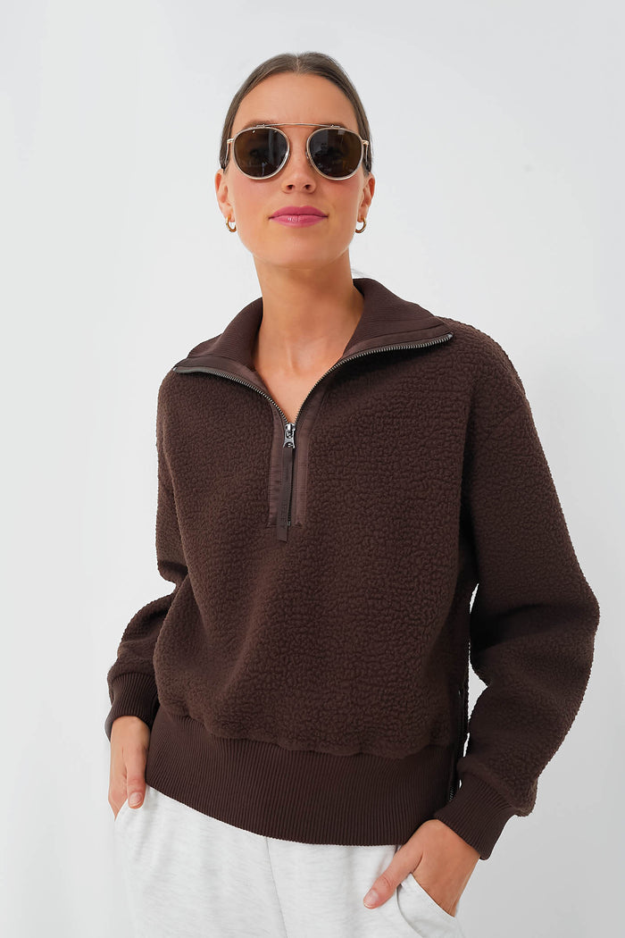 A woman wears sunglasses, light-colored pants, and a Varley Dark Truffle Roselle Half Zip Fleece crafted from recycled polyester fleece.