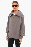 A woman in black sunglasses, gold hoop earrings, and a Varley half zip pullover with a wide collar and balloon sleeves stands confidently, highlighting Varley's signature blend of performance and style.