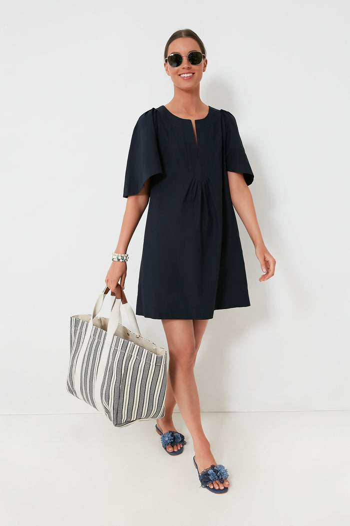 A person in a Pomander Place black mini dress with flutter sleeves, sunglasses, and slide sandals carries a large striped tote bag.