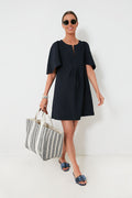 A person in a Pomander Place black mini dress with flutter sleeves, sunglasses, and slide sandals carries a large striped tote bag.