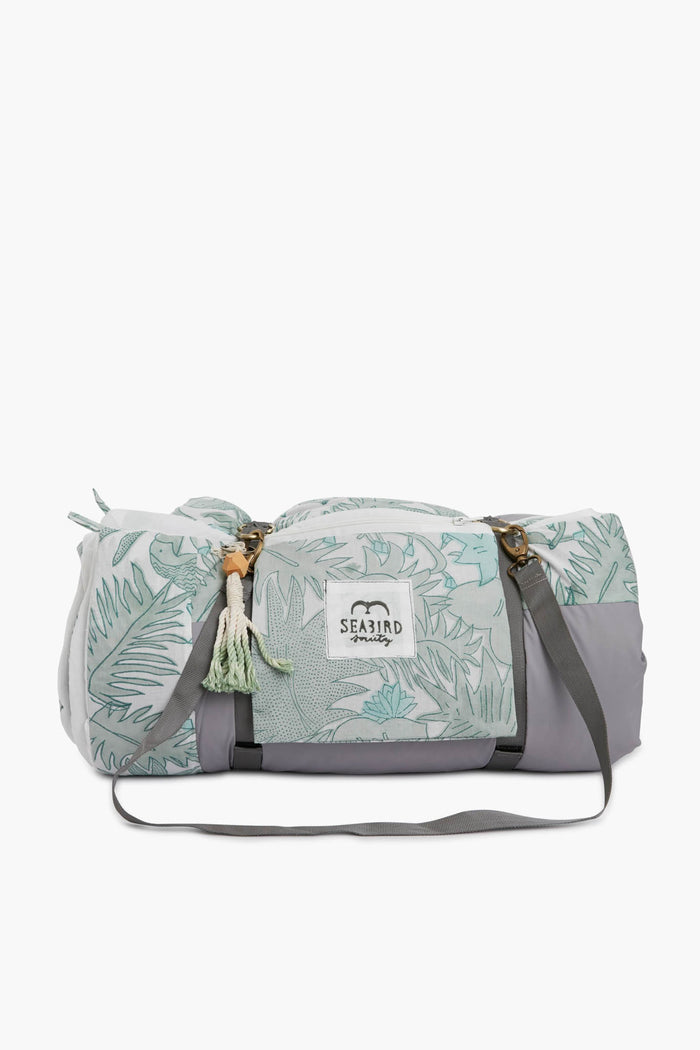 A gray and mint green Seabird Society duffel bag with leaf patterns, front pocket, shoulder strap, carrying handles, and logo patch—perfect for carrying your outdoor quilted blanket.